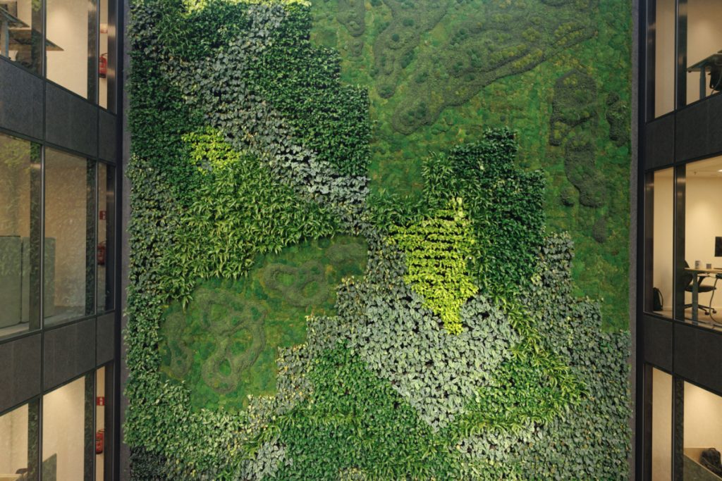 Espectacular muro verde (jardín vertical) interior de gran altura que cubre una pared completa de un atrio o lobby de varios pisos. El muro vegetal presenta un diseño con texturas y patrones variados, utilizando diferentes tonos de verde, incluyendo musgo, helechos, y otras plantas de hoja pequeña, con algunas áreas de vegetación más clara que forman contornos abstractos. El muro se extiende desde el suelo hasta el techo, flanqueado por la fachada de vidrio y acero negro de las oficinas o habitaciones en los pisos superiores. El propósito de la imagen es mostrar un diseño de interior a gran escala que utiliza un jardín vertical masivo para crear un punto focal natural, mejorar la estética, la acústica y la calidad del aire en un entorno corporativo de lujo.