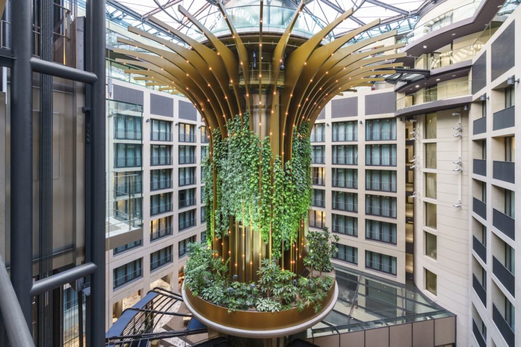Vista de un atrio de hotel o edificio de oficinas de varios pisos. La característica central es una impresionante escultura o jardín vertical suspendido que tiene forma de flor o palmera estilizada, con pétalos dorados grandes y un tronco central cilíndrico. El tronco está cubierto por plantas trepadoras y colgantes (jardín vertical), y la base inferior alberga un parterre de plantas más grandes, todo iluminado con luces focales. La estructura se eleva en el centro del atrio, el cual está rodeado por múltiples pisos de habitaciones o suites con fachadas grises y ventanales uniformes. El techo del atrio es un lucernario de vidrio y metal. El propósito de la imagen es mostrar un diseño de interior espectacular que combina arquitectura moderna y un elemento biofílico monumental como punto focal, enfatizando el lujo, la innovación y la conexión con la naturaleza en un entorno de gran escala.